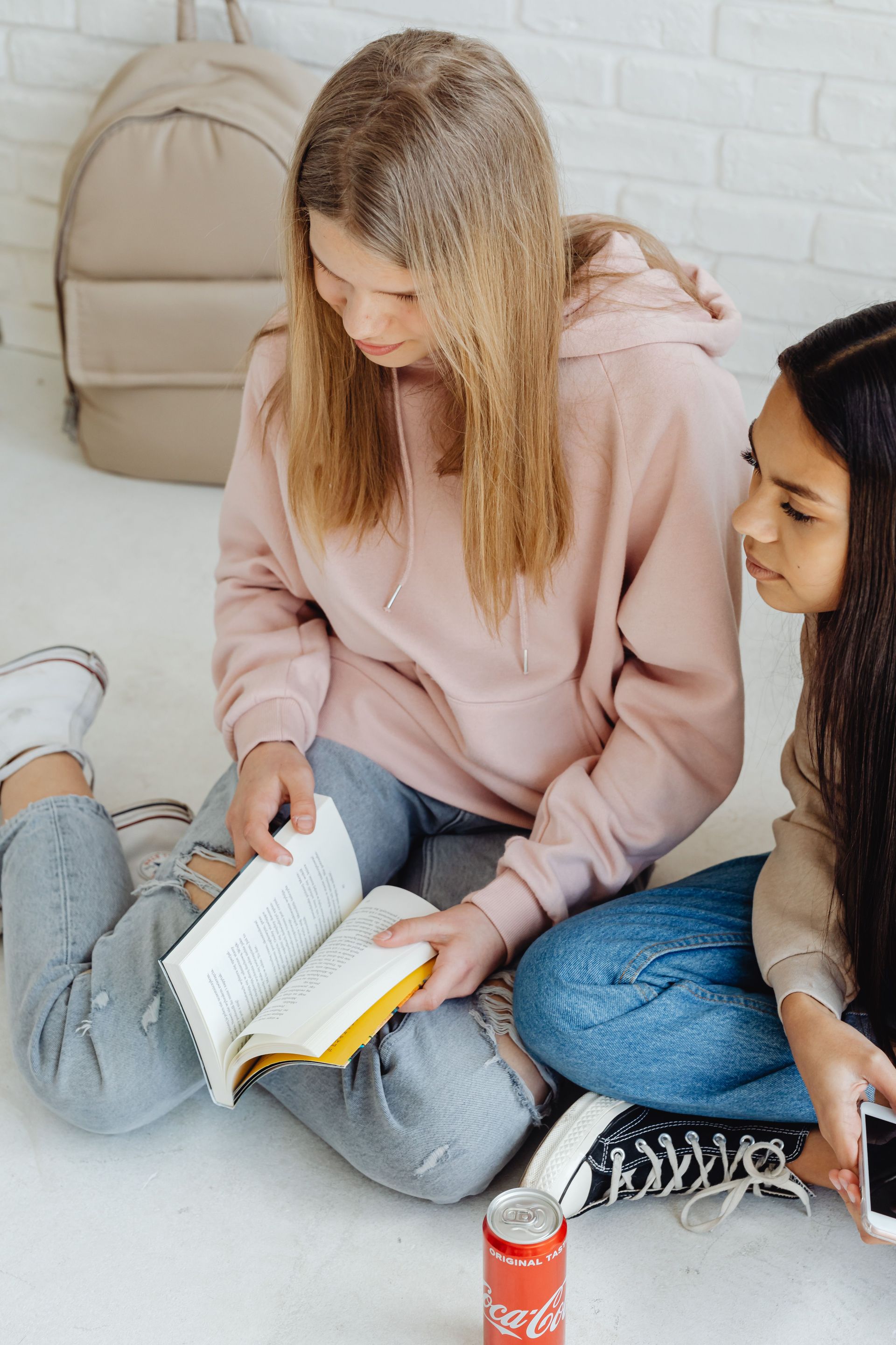 Two teenagers sitting on the floor, reading and enjoying a soda. Casual indoor setting with a backpack.
